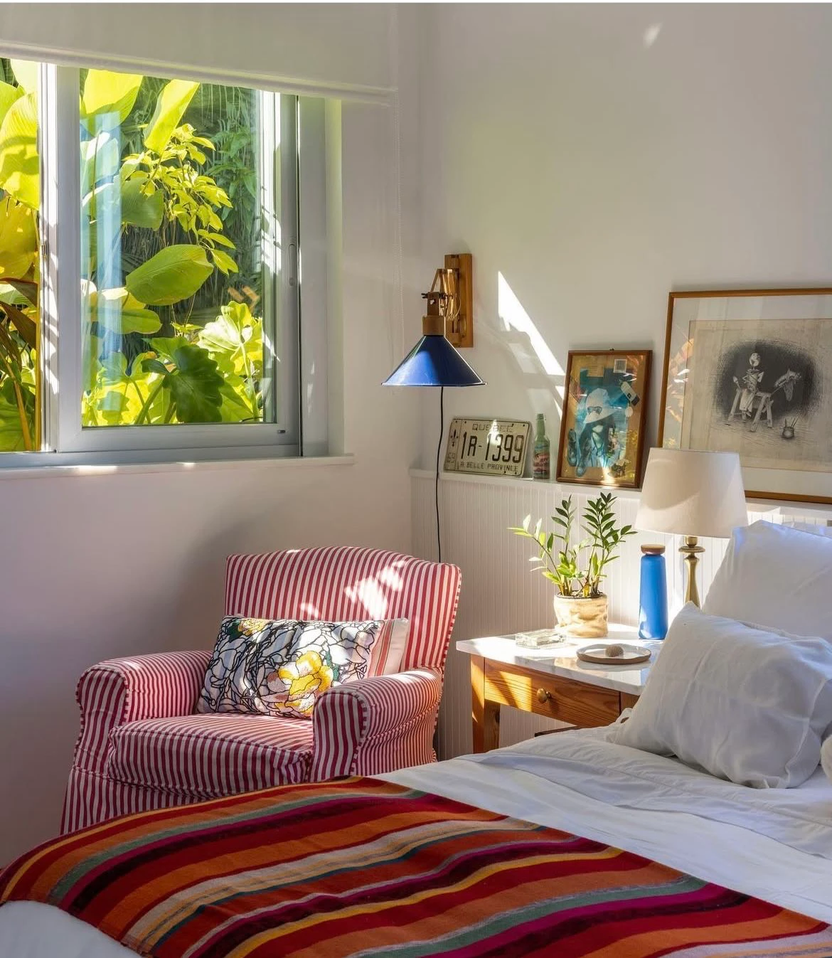 Quarto com poltrona listrada de vermelho, luminária azul de parede, quadros na prateleira e manta colorida na cama, com janela para plantas tropicais