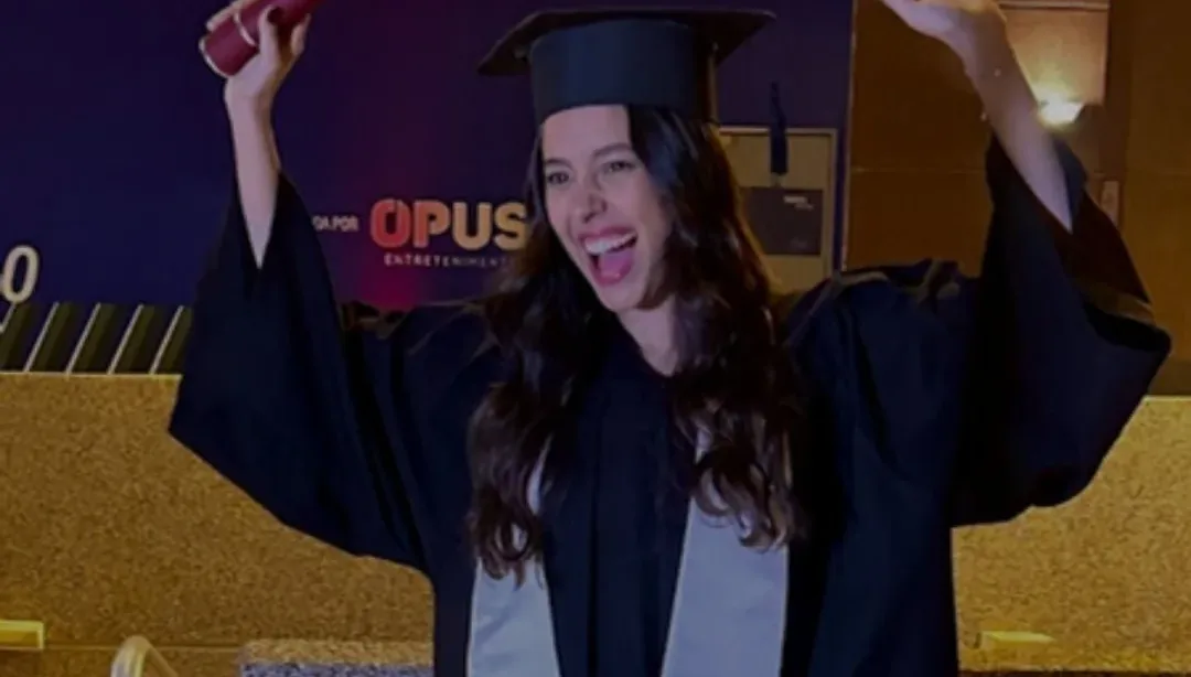 Mariana de beca e capelo, sorrindo e levantando o diploma na formatura da ESPM