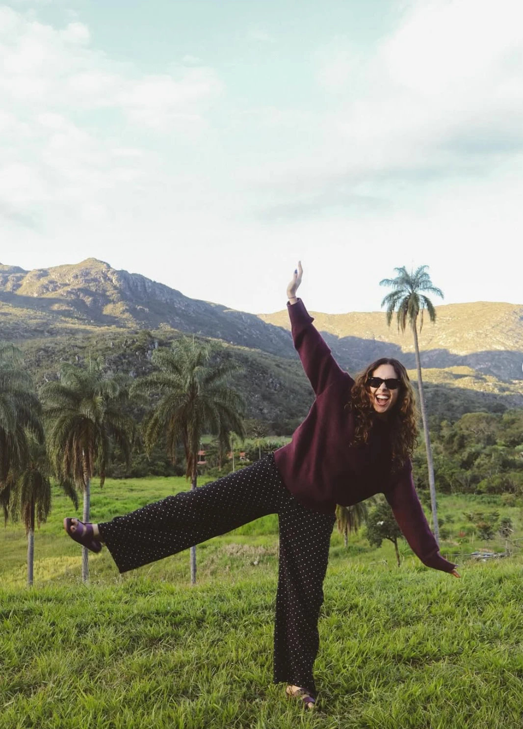Mariana de óculos escuros, suéter vinho e calça de bolinhas, equilibrada numa perna só e de braços abertos num campo verde com palmeiras e serra ao fundo